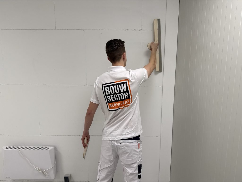 Stukadoor van Bouwsector Nederland trekt een wand van kalkzandsteen recht met een brede spaan, naast een elektrische kachel