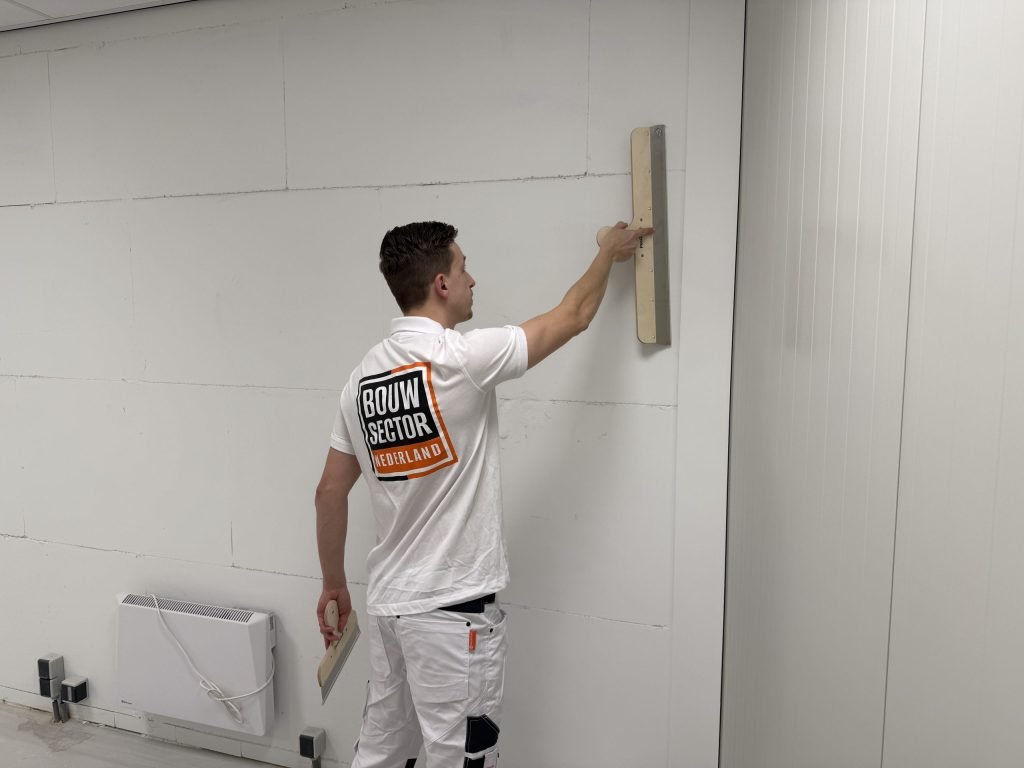 Stukadoor van Bouwsector Nederland in wit werkpak trekt met een lange spaan een wand strak, met een spaan in zijn andere hand