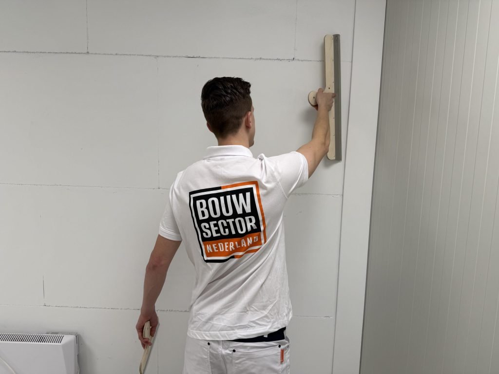 Stukadoor van Bouwsector Nederland met logo op de rug werkt met een lange spaan hoog op een wand