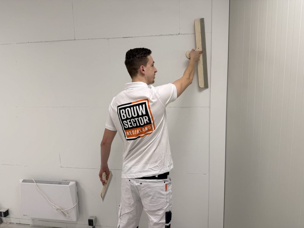 Stukadoor van Bouwsector Nederland in wit werkpak houdt een lange spaan tegen de wand en een kleine spaan in zijn andere hand