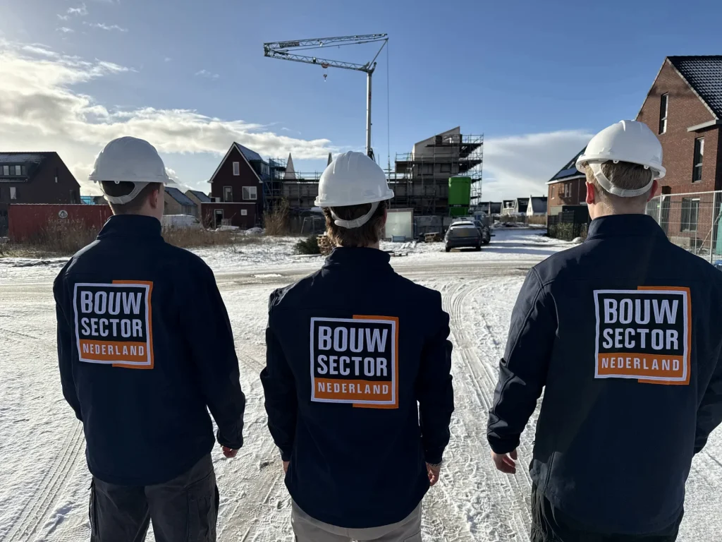 Drie vakmensen van Bouwsector Nederland met witte helm en bedrijfsjas kijken over een besneeuwde nieuwbouwstraat