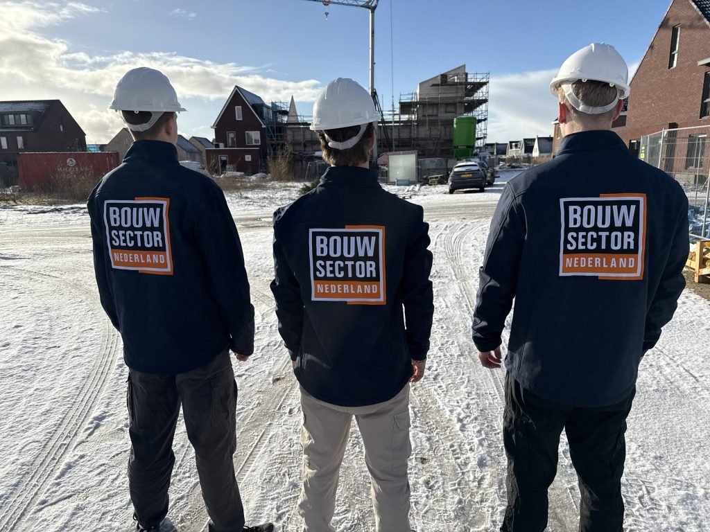 Drie vakmensen van Bouwsector Nederland met witte helm en bedrijfsjas lopen over een besneeuwde bouwplaats met nieuwe woningen